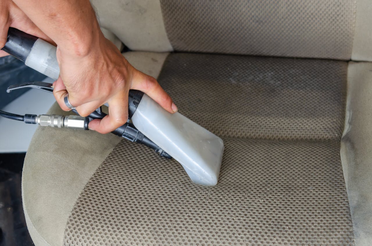 why-choose-us-03 Hands using a vacuum nozzle to deeply clean a fabric car seat, removing dirt and stains.
