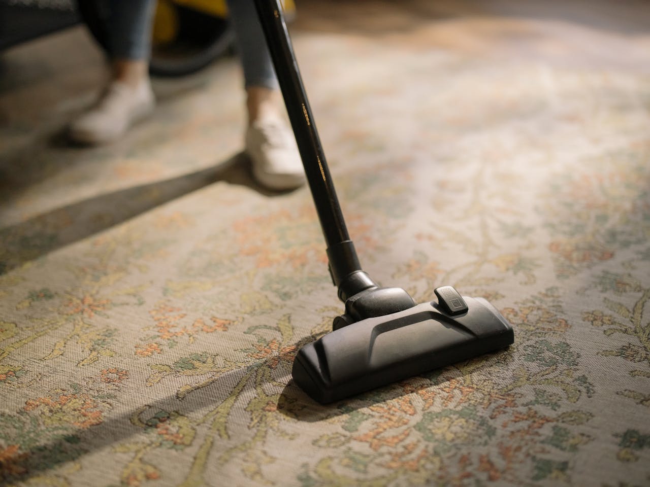 hero-01 Close-up of a vacuum cleaner on a patterned carpet in a sunlit room, capturing a moment of household cleaning.